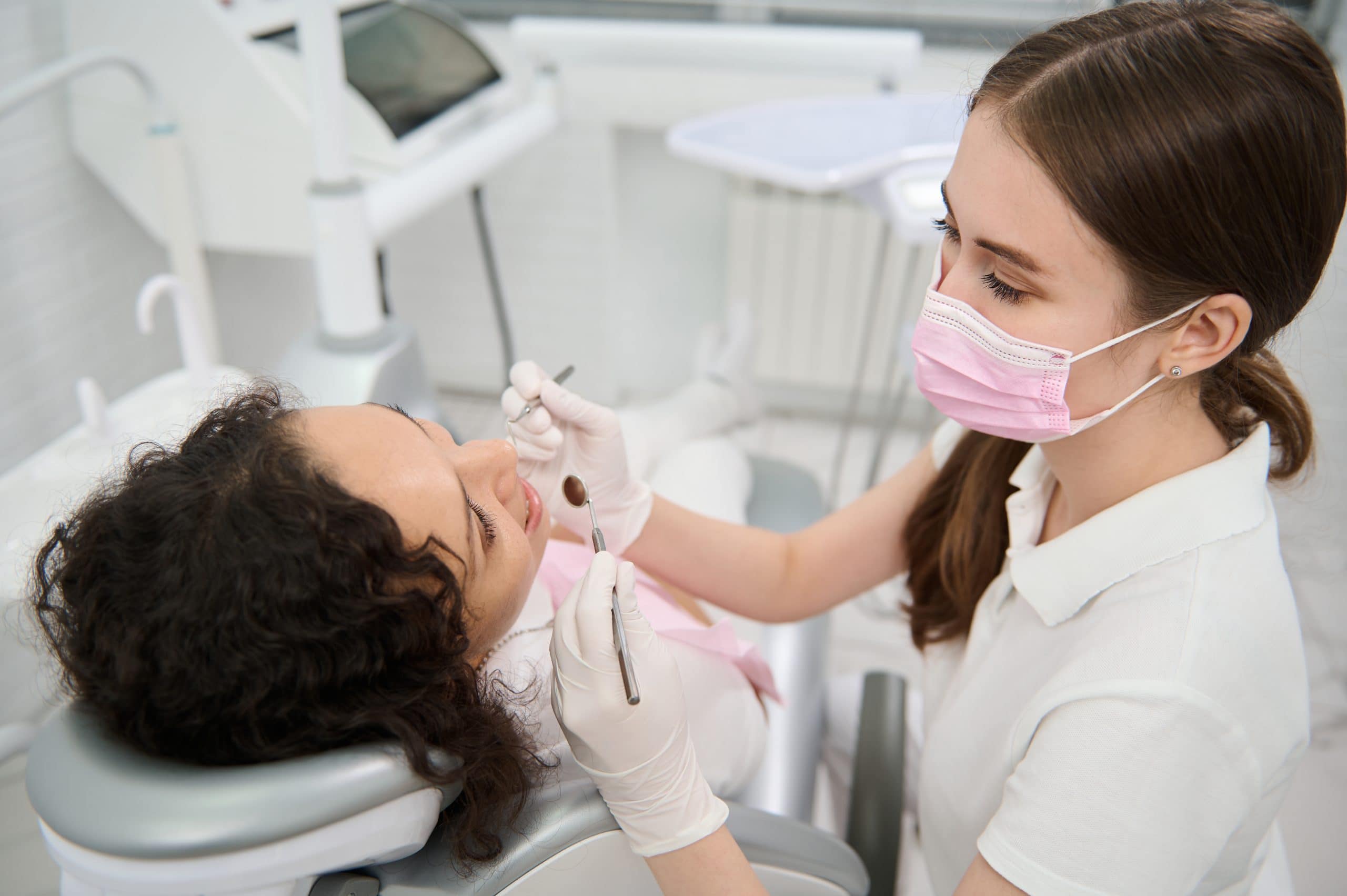 dentist hygienist in medical protective mask examining the oral cavity of a female patient with sterile stainless steel dental mirror and probe in dentistry clinic during planned medical check up